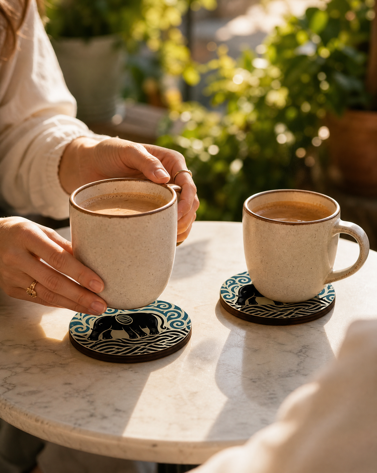 Two beige speckled ceramic mugs with dark brown rims sit on matching coasters on a white marble table. One mug is being held by hands, and the background shows a softly blurred outdoor patio setting with green plants and warm sunlight.